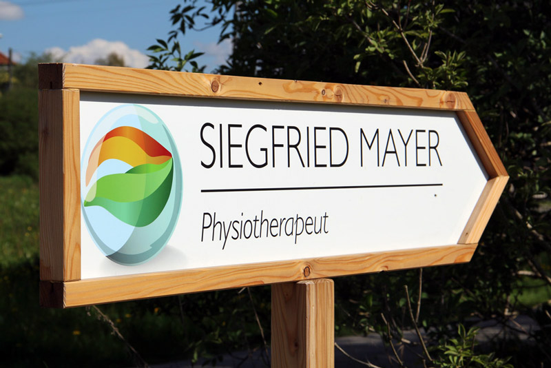 große Holztafel in Pfeilform. Text-Inhalt: Siegfreid Mayer, Physiotherapeut